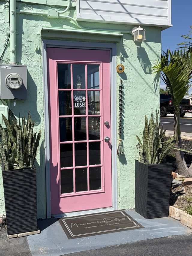 The shop’s pink door on a mint green wall, with a Mariann’s Boutique doormat and two potted plants